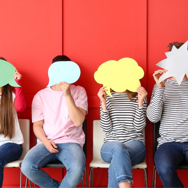 Četri cilvēki sēž pie sarkanas sienas, aizklājot savas sejas ar dažādu krāsu runas un domu burbuļiem – zaļu, gaiši zilu, dzeltenu un baltu.