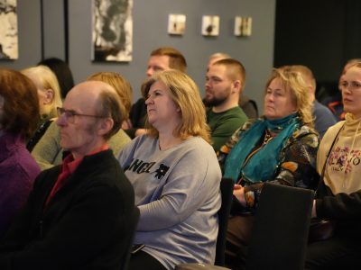 Daudzveidīga cilvēku grupa sēž auditorijā, skatoties uz priekšu.