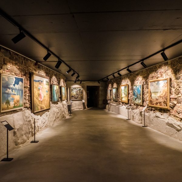 Plaša gleznu izstāde šaurā, garenā telpā ar neapstrādātām akmens sienām un tumšu griestiem, kur gleznas izgaismo prožektori.