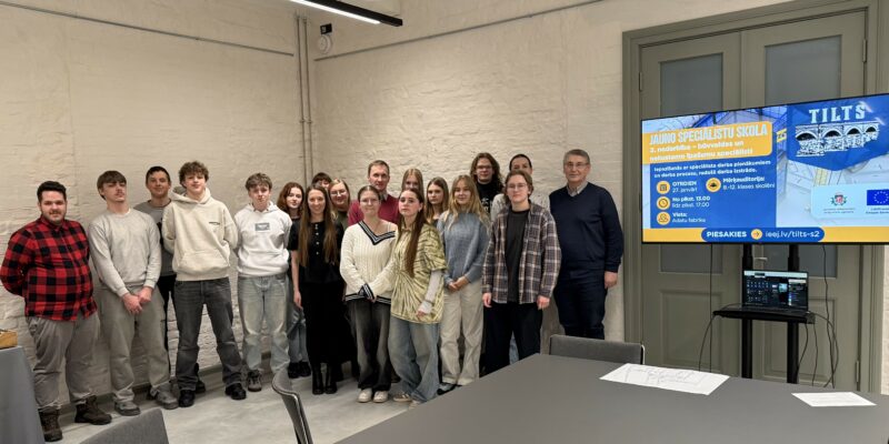 Aptuveni divdesmit jaunieši un viens pieaugušais vīrietis pozē grupas fotogrāfijai gaišā telpā. Aiz viņiem ir liels ekrāns ar informāciju par "JAUNO SPECIĀLISTU SKOLU" un "2. nodarbība – būvvaldes un nekustamo īpašumu speciālisti". Teksts uz ekrāna min: "OTRDIEN 27. janvārī", "No plkst. 13.00 līdz plkst. 17.00", "Vieta: Adatu fabrika", "Mērķauditorija: 8.-12. klases skolēni" un "PIESAKIES ieeja.lv/tilts-s2".