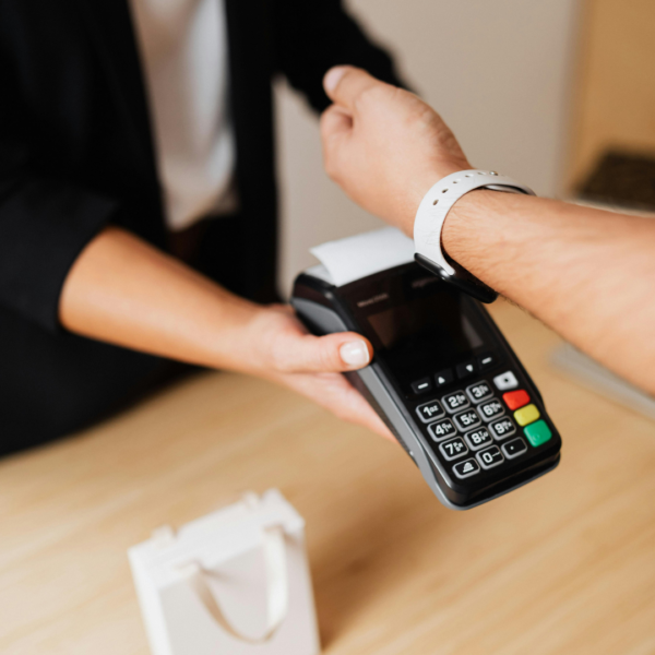 Tuvplānā redzams, kā cilvēks ar baltu viedpulksteni uz rokas veic bezkontakta maksājumu, pieliekot to pie melna POS termināļa, ko tur cits cilvēks.