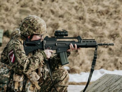 Notupies karavīrs kamuflāžas uniformā ar Latvijas karoga plāksteri un ķiveri mērķē ar šauteni ar optisko tēmekli, valkājot dzirdes aizsargus.