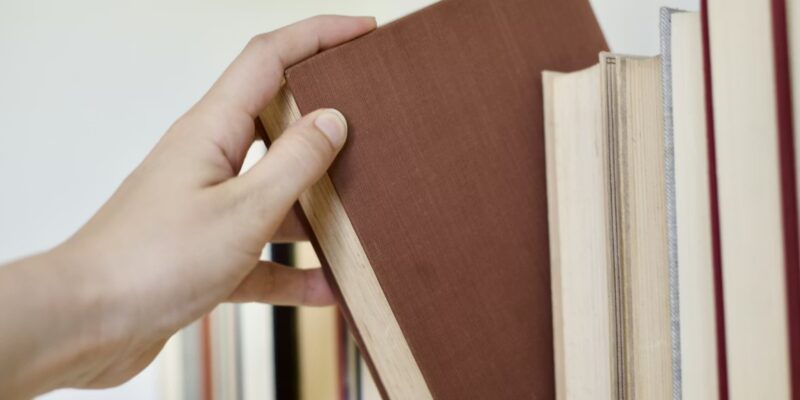 Cilvēka roka izvelk brūnu grāmatu no grāmatu plaukta, kurā redzamas arī citas grāmatas.